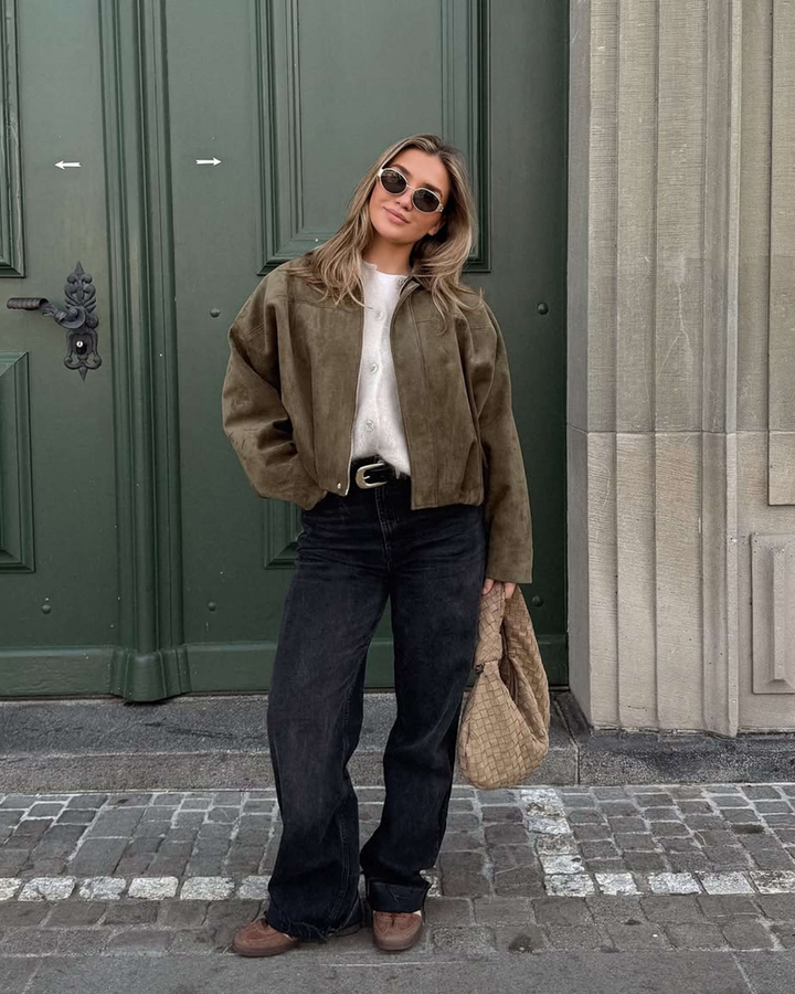 Woman in a brown jacket and black jeans standing in front of a green door.