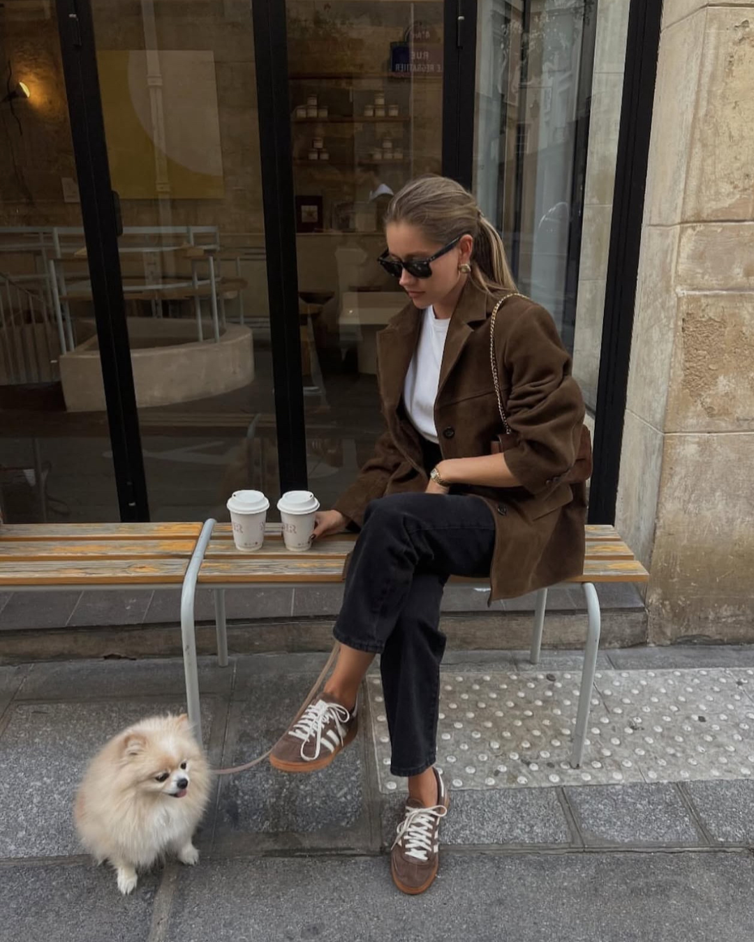 Woman sitting on a bench with a dog, holding two coffee cups in an urban setting.