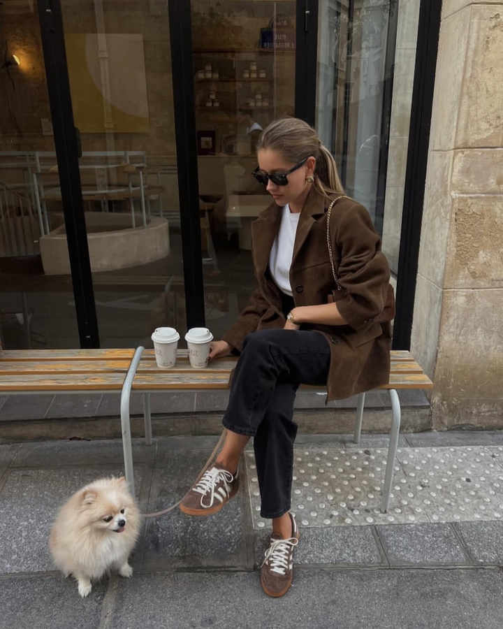 Woman sitting on a bench with a dog, holding two coffee cups in an urban setting.