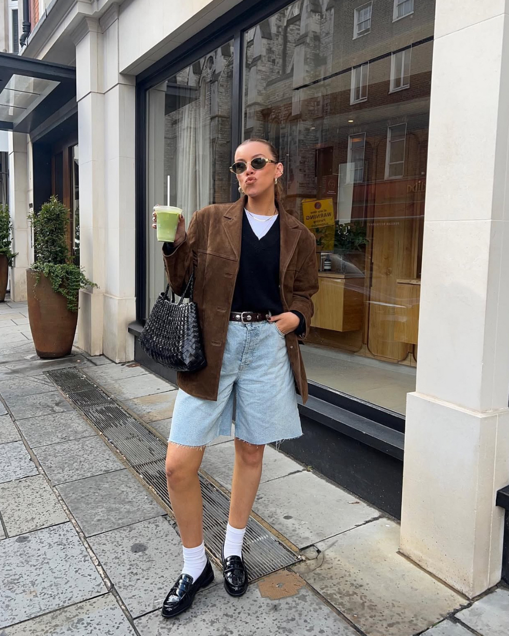Woman in stylish outfit standing on a city street, holding a drink.