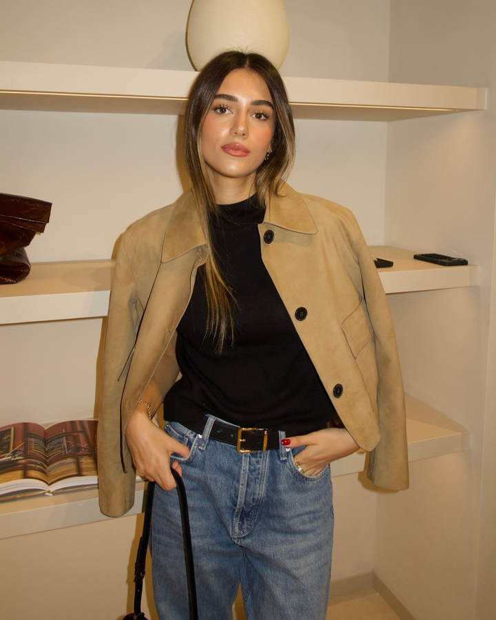 Woman wearing a beige coat, black top, and blue jeans indoors.