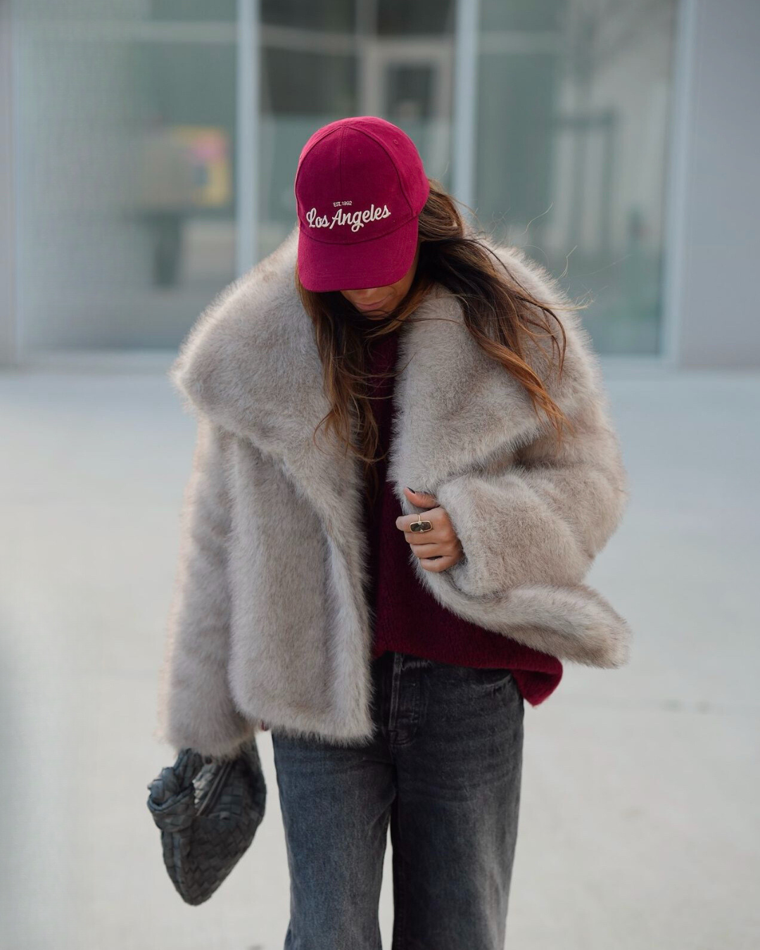 Person wearing a fluffy beige coat, maroon sweater, gray jeans, and pink 'Los Angeles' cap.