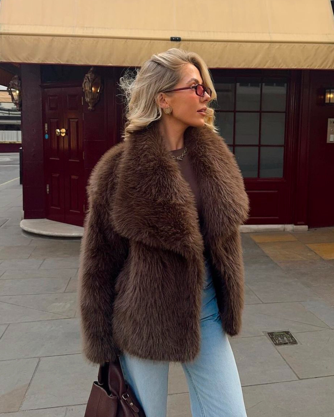 Woman wearing a brown fur coat and light blue jeans standing in front of a building entrance.