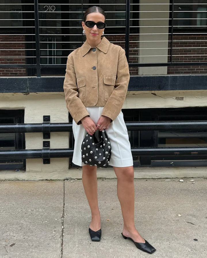Woman wearing a beige jacket and white skirt, holding a polka dot bag, standing in front of a building.