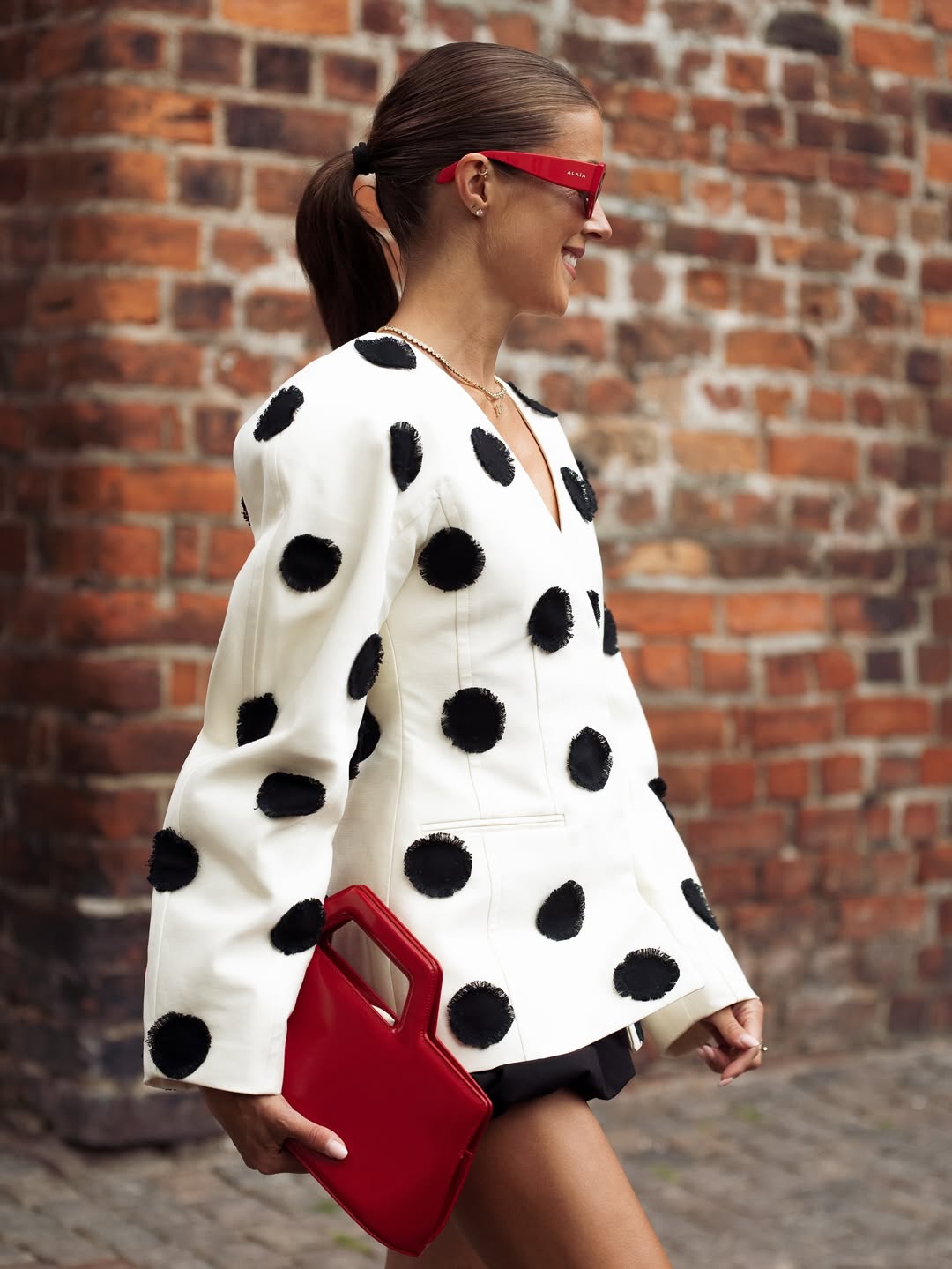 Woman wearing a white polka dot cardigan and red handbag against a brick wall.