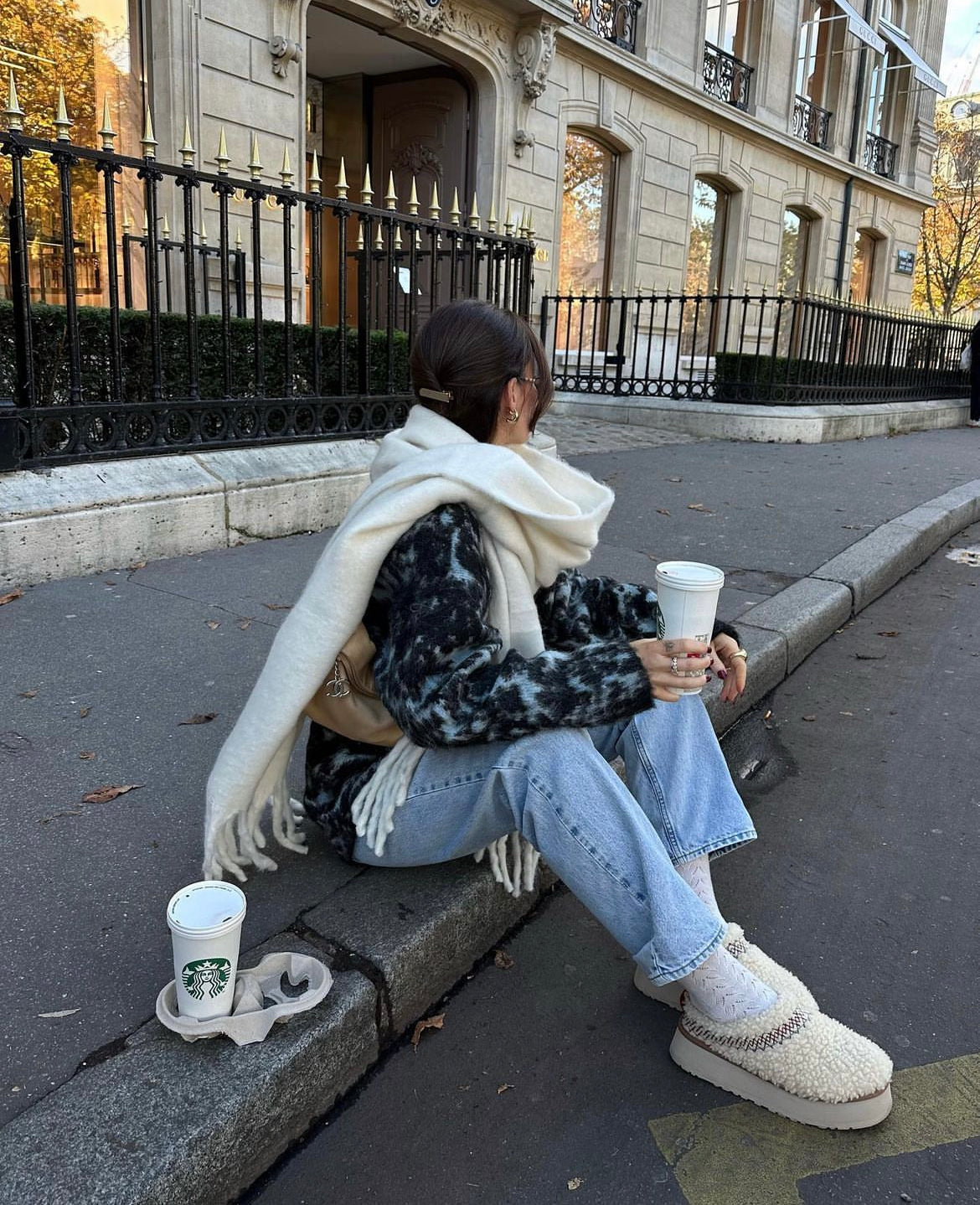 Person sitting on a sidewalk with a Starbucks cup, wearing a scarf and jeans.