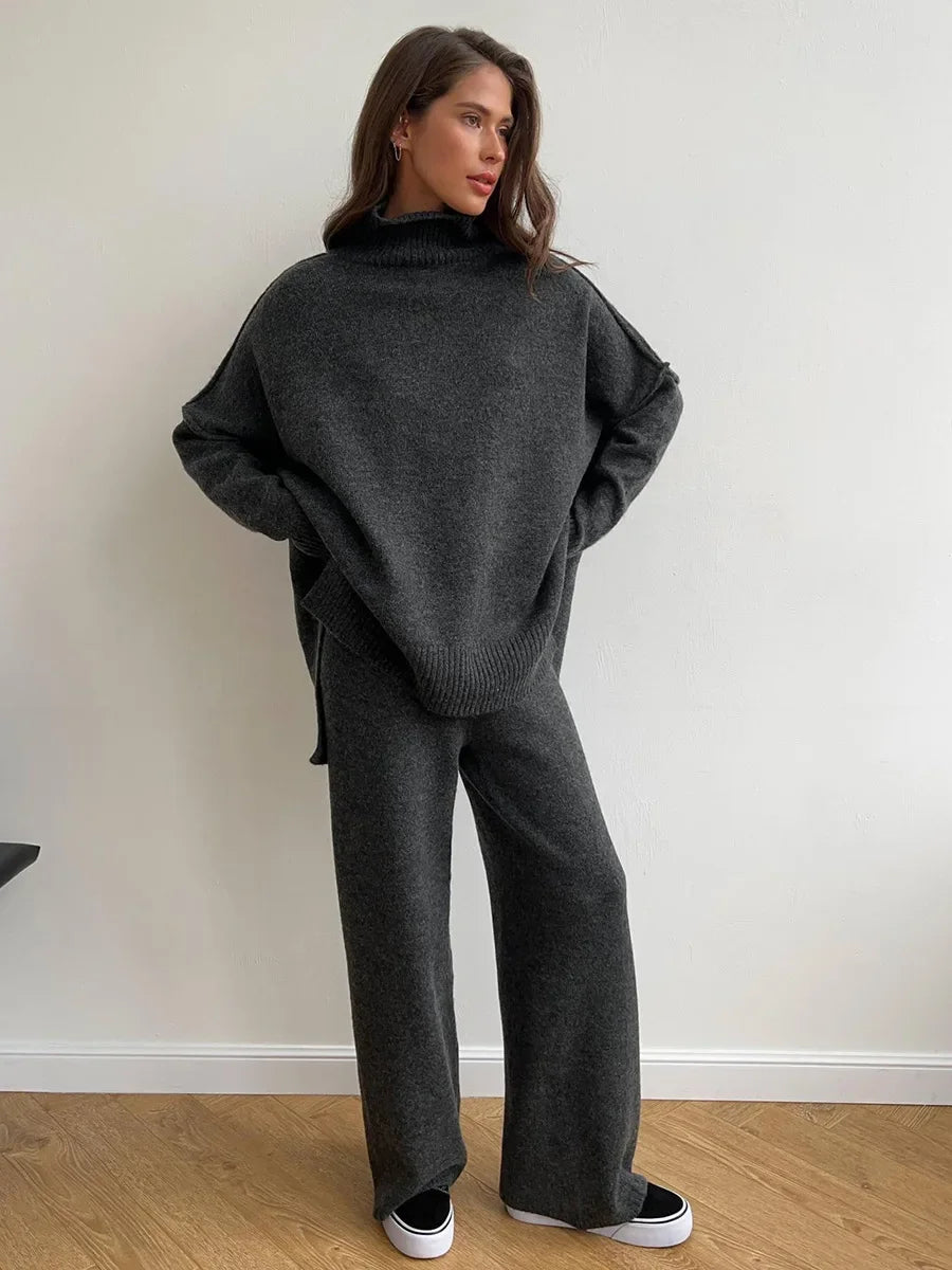 Woman wearing a dark gray sweater and pants against a white wall.