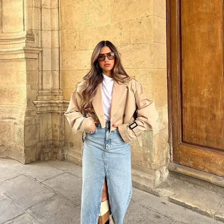 Woman in beige jacket and blue jeans standing in front of a stone wall.