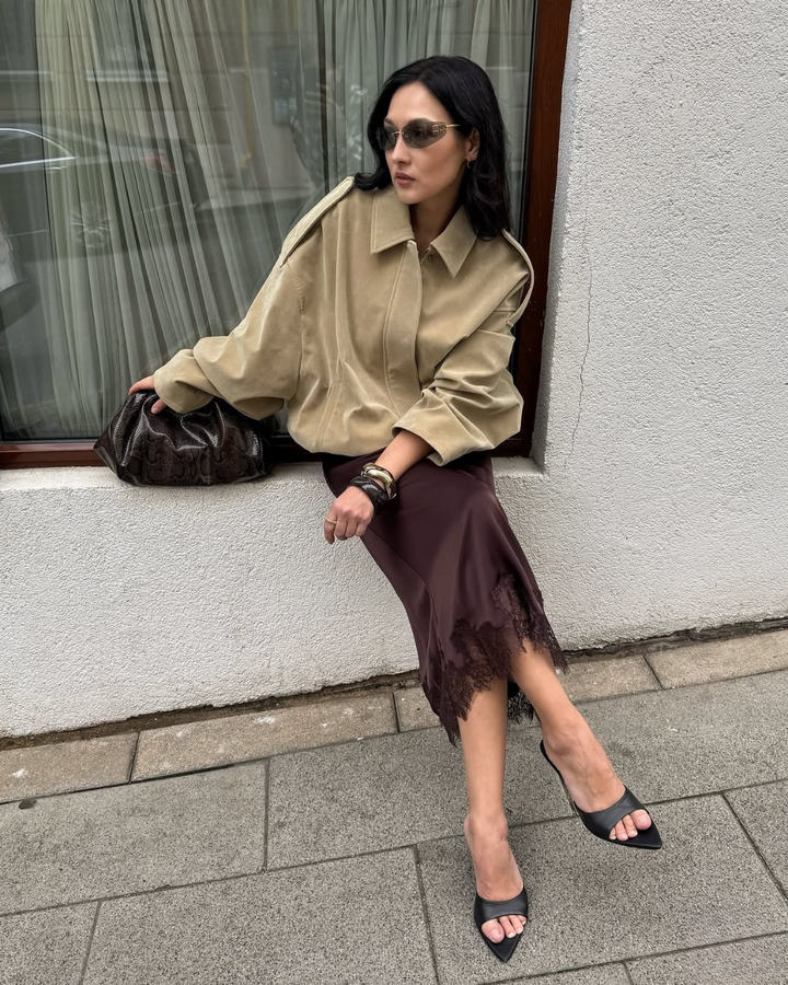 Woman in beige shirt and brown skirt sitting on a ledge outdoors.