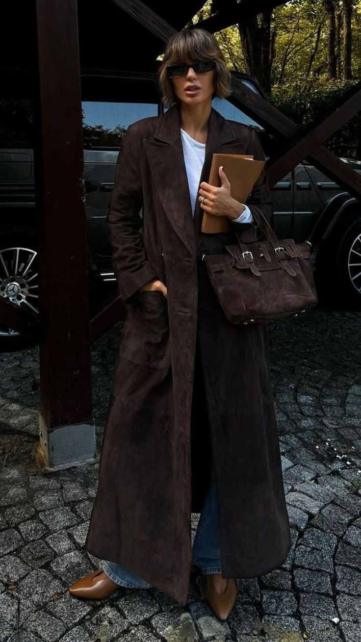 Person wearing a long brown coat holding a brown bag and a folder, standing on a cobblestone street.