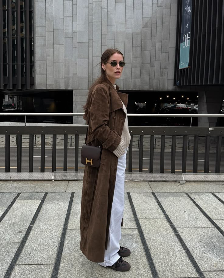 Woman wearing a brown coat and sunglasses, standing on a city street.