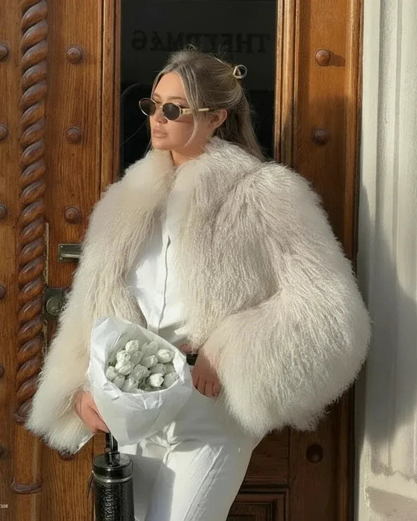 Woman in a white fur coat and sunglasses holding flowers, standing in front of a wooden door.