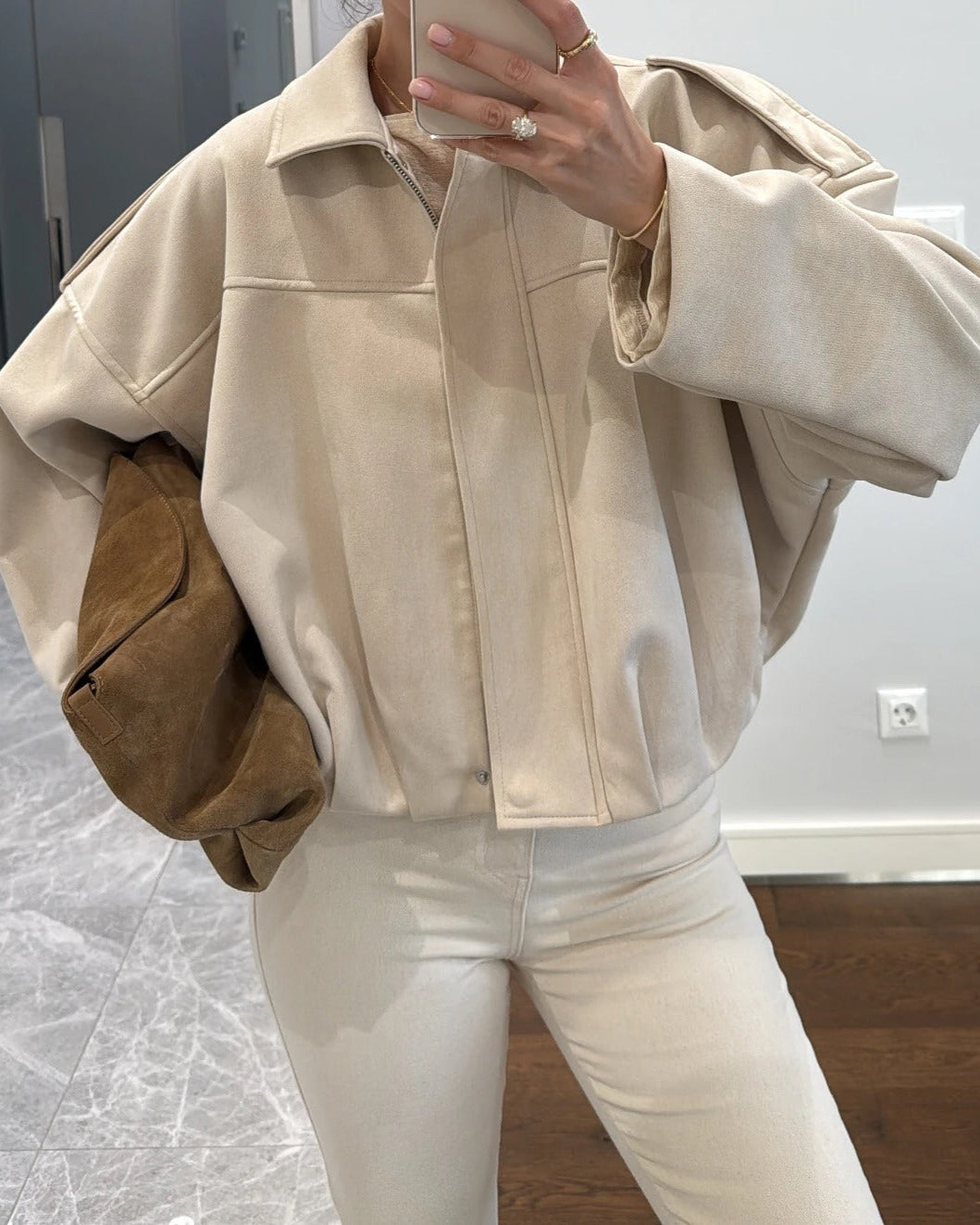 Person wearing a beige jacket and white pants taking a mirror selfie.