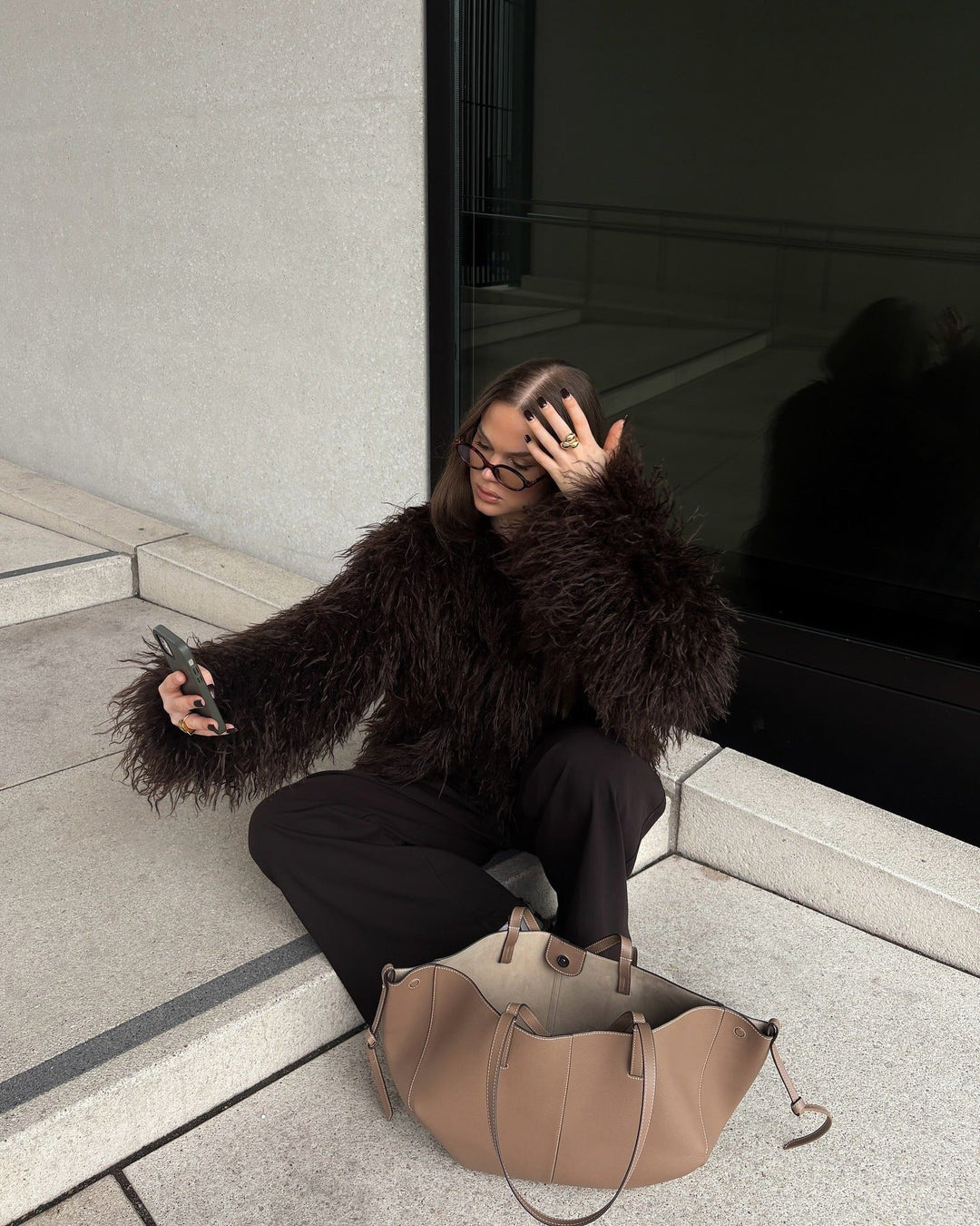 Person sitting on steps wearing a dark fur coat with a beige handbag nearby.
