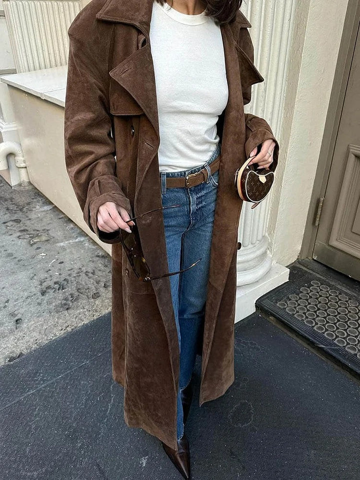Person wearing a brown coat, white shirt, and blue jeans standing on a sidewalk.