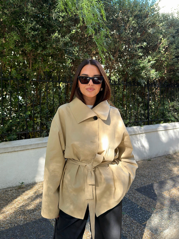 Woman wearing a beige trench coat and sunglasses outdoors with greenery in the background