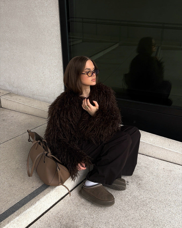 Person sitting on a step wearing a dark fur coat and glasses, with a reflective surface behind them.
