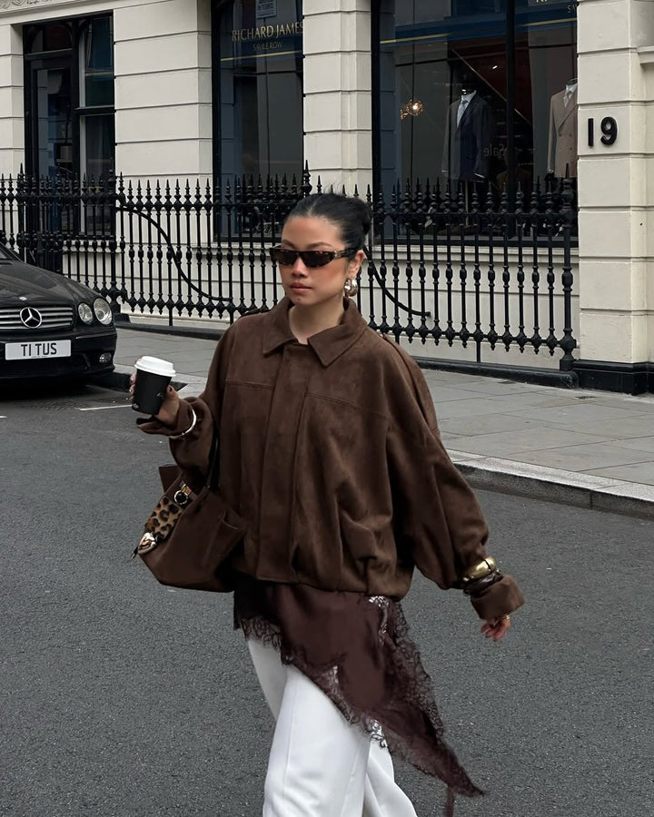 Woman in a brown jacket and white pants walking on a street with a building and car in the background.