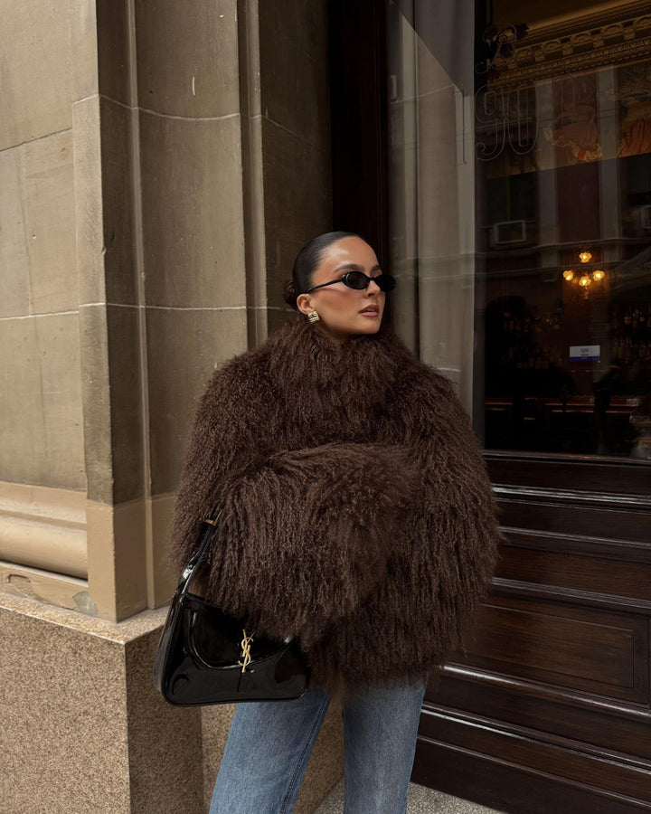 Woman wearing a brown fur coat and black sunglasses, standing in front of a building entrance.